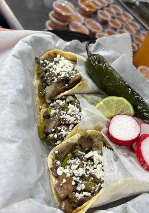 Two tacos filled with meat and vegetables, topped with cheese, served with a jalapeño, lime, and radishes on the side.