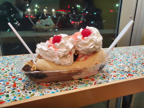 A banana split topped with whipped cream and cherries, served in a clear dish on a colorful countertop.