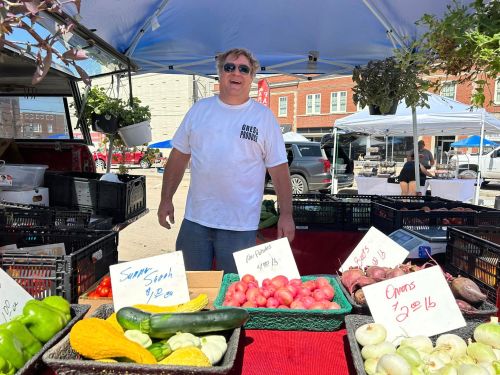 A smiling vendor stands at a market stall with various fresh vegetables and price signs.