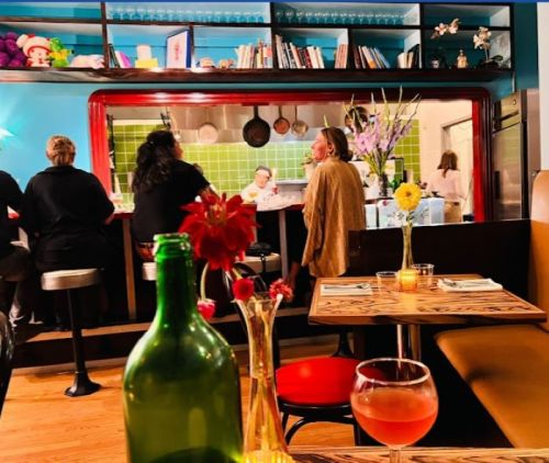A cozy restaurant scene with patrons at the bar, colorful decor, and a table set with drinks and flowers in the foreground.