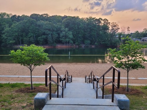 Steps leading down to a sandy beach by a calm lake, surrounded by trees under a colorful sunset sky.