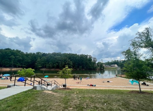 A sandy beach by a lake, with people relaxing under umbrellas and trees, set against a cloudy sky.