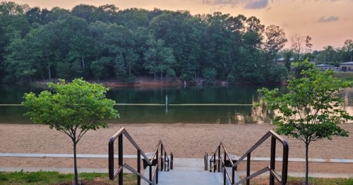 A serene view of a sandy beach leading to a calm lake, surrounded by trees under a pastel sky.