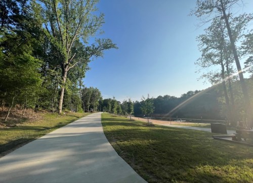 A paved path winds through a green park with trees, leading to a sandy area in the distance under a clear blue sky.