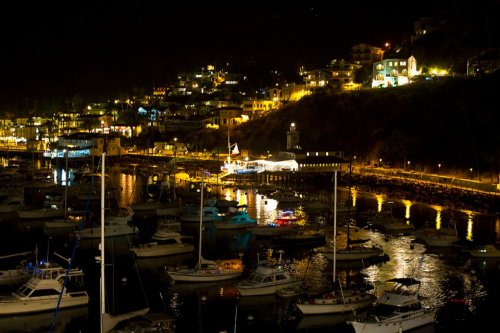 A nighttime view of a harbor filled with boats, illuminated by lights from nearby buildings and reflections on the water.