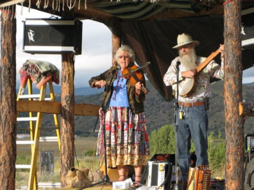 A woman plays the violin while a man with a banjo performs on a rustic stage in a scenic outdoor setting.