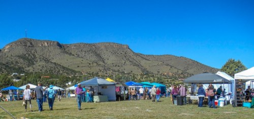 A vibrant outdoor market with tents and people, set against a backdrop of mountains under a clear blue sky.