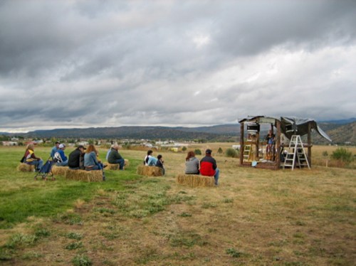 A group of people sitting on hay bales, watching a presentation at an outdoor venue under a cloudy sky.