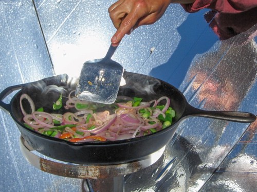 A hand stirs sautéing onions and peppers in a cast iron skillet over a heat source, with steam rising.