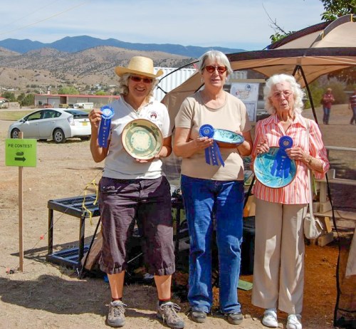 Three women hold blue ribbons and pie plates, smiling at an outdoor event with mountains in the background.