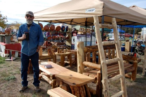 A bearded man stands by rustic wooden furniture at an outdoor market, holding a coffee cup under a tent.