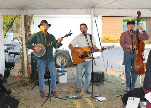 Three musicians perform under a tent, playing banjo, guitar, and double bass at an outdoor event.