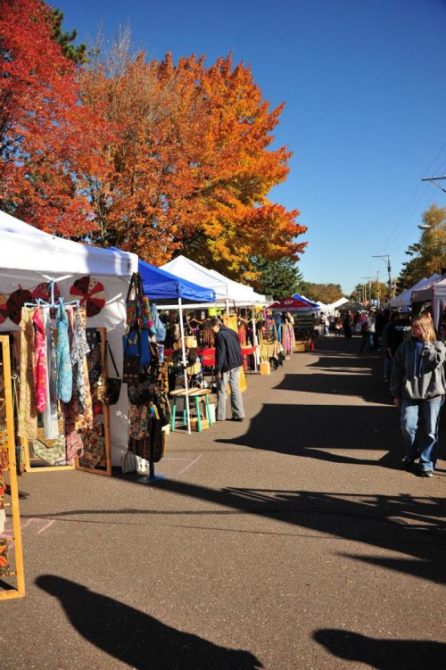 A vibrant outdoor market with colorful tents, autumn trees, and people browsing various stalls.