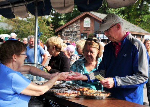A man pays for baked goods at a market stall while two women watch, surrounded by a crowd in the background.