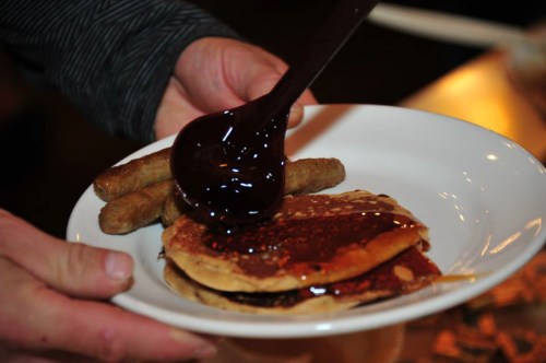 A person drizzles chocolate sauce over pancakes on a plate, accompanied by sausage links.