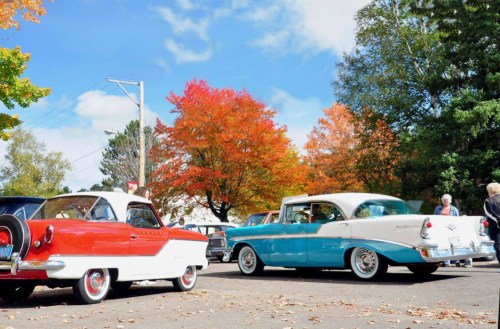 Two classic cars, one red and white and the other turquoise and white, parked under colorful autumn trees.