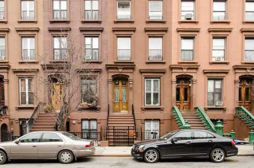 Brownstone buildings with green and brown steps, parked cars on the street, and a bare tree in front.