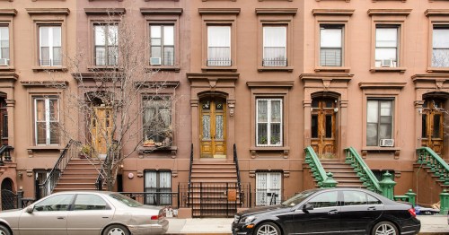 A row of brownstone buildings with steps, windows, and parked cars on a city street.