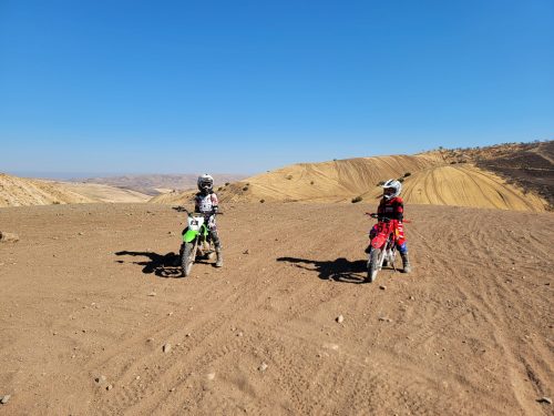 Two kids on dirt bikes in a sandy, hilly landscape under a clear blue sky.