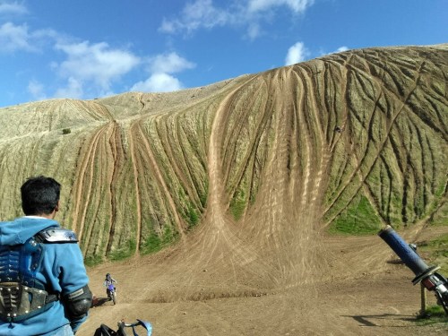 A person stands on a dirt path, looking at a steep, rutted hill with tire tracks and blue sky above.