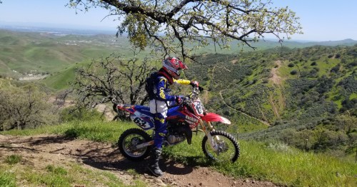 A motocross rider in gear stands beside a dirt bike on a scenic hillside with green hills in the background.