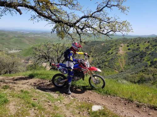 A motocross rider in gear stands beside a dirt bike on a scenic hillside with green hills and blue sky in the background.