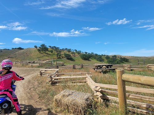 A person on a dirt bike rides along a trail with wooden fences and green hills under a blue sky.