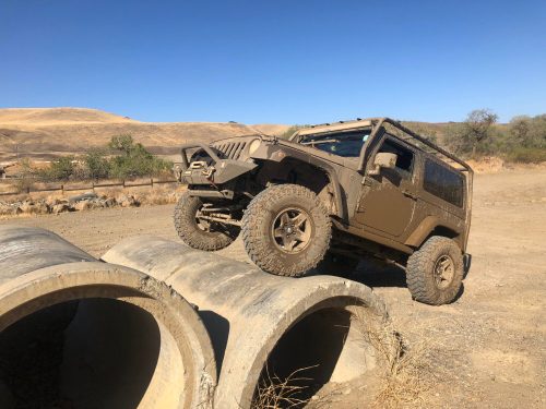 A muddy off-road vehicle perched on concrete pipes in a dry, hilly landscape under a clear blue sky.