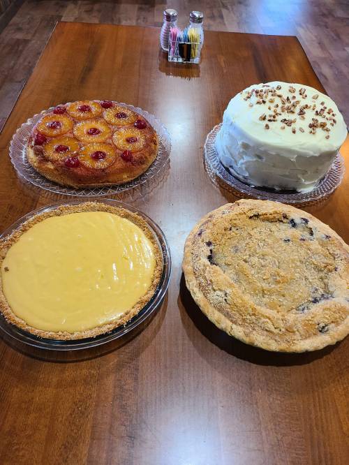 Four desserts on a wooden table: a fruit-topped cake, a white frosted cake, a lemon pie, and a blueberry crumble pie.