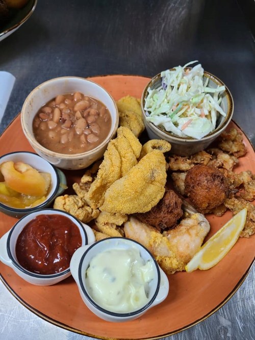 A colorful plate featuring fried fish, coleslaw, baked beans, and various dipping sauces, garnished with lemon.