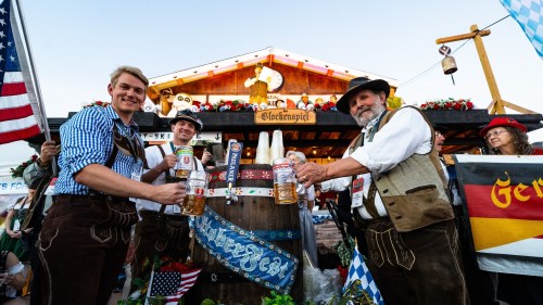 Three men in traditional Bavarian attire raise beer mugs at a festive outdoor event, with flags and decorations in the background.
