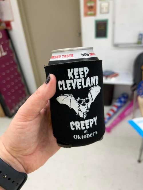 A hand holding a black can cooler with the text "Keep Cleveland Creepy" and a bat graphic, promoting Oktoberfest.