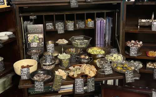 A display of various crystals and stones in bowls, labeled with names, on a wooden shelf with books and decorative items.