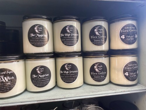 A shelf displaying various scented candles in jars, labeled with names like "The Magician" and "Flora Gotica."
