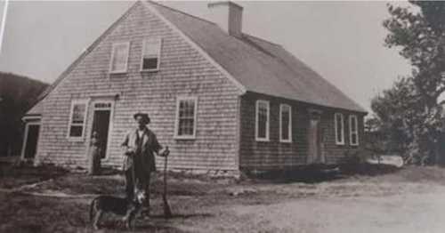 A black and white photo of a man with a cane standing beside a dog in front of a wooden house.