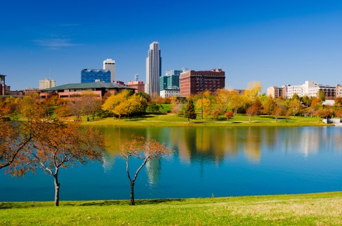City skyline reflected in a calm lake, surrounded by colorful autumn trees and clear blue skies.