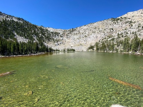 A clear, tranquil lake surrounded by rocky mountains and lush green trees under a bright blue sky.