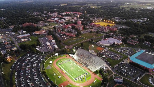 Aerial view of a stadium at dusk, surrounded by a bustling campus and parking lot, with lights illuminating the field.