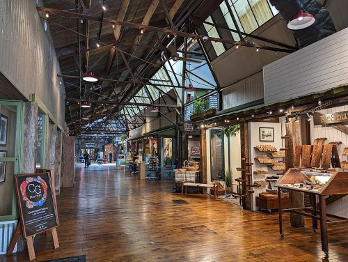 A spacious indoor market with wooden floors, shops on either side, and warm lighting creating a cozy atmosphere.