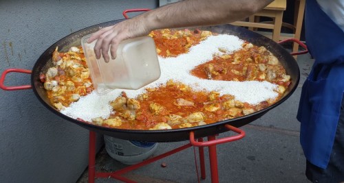 A person pours rice into a large pan of simmering chicken and sauce outdoors.