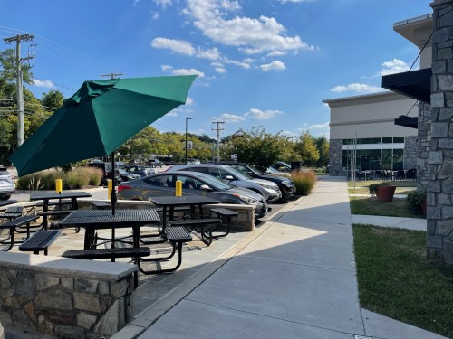 A sunny outdoor area with a green umbrella, picnic tables, and parked cars beside a modern building.