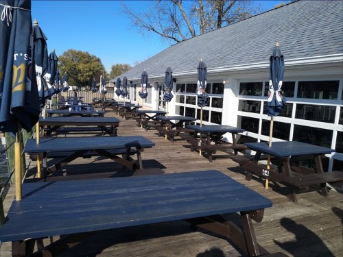 Outdoor dining area with wooden tables and blue umbrellas, set along a waterfront on a sunny day.