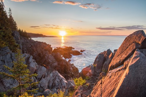 Sunset over the ocean, with rocky cliffs and trees framing the scene, casting warm colors across the sky and water.
