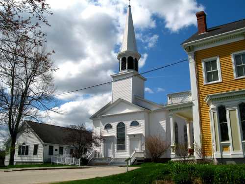 A white church with a tall steeple beside a yellow building, set against a blue sky with fluffy clouds.