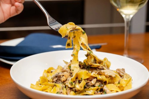 A fork lifts creamy pasta with mushrooms from a white bowl, accompanied by a glass of white wine.
