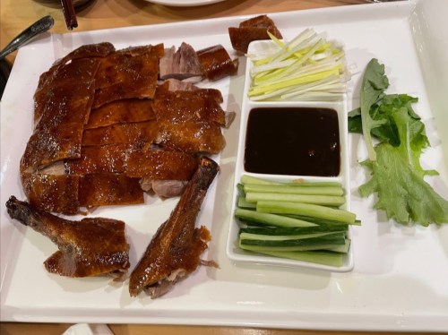 A platter of sliced roasted duck served with green onions, cucumber sticks, lettuce, and a small bowl of sauce.