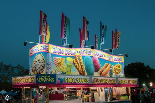 Colorful food stand at a fair featuring corn dogs, cotton candy, funnel cakes, and more under a twilight sky.