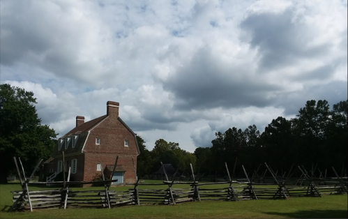 A historic brick house beside a wooden fence, set against a backdrop of cloudy skies and green trees.