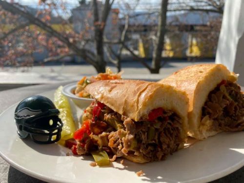 A close-up of a sandwich filled with meat and peppers, accompanied by a pickle and a small football helmet.
