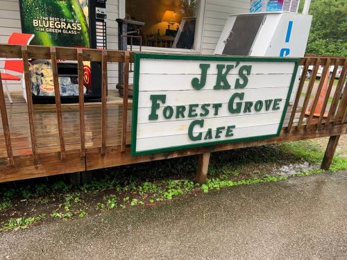 Sign for JKS Forest Grove Cafe on a wooden deck, with a rainy outdoor setting in the background.